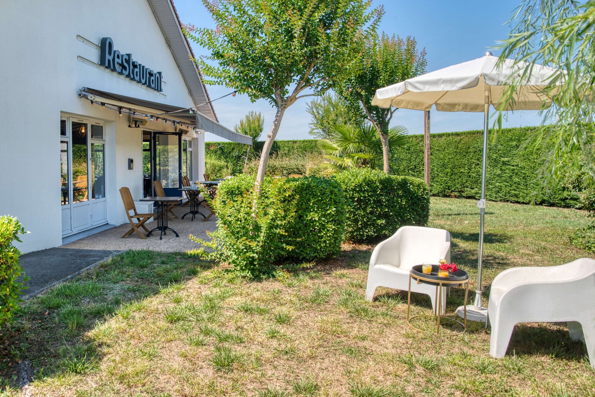 Jardin de l'hôtel avec parasol, fauteuils et terrasse du restaurant