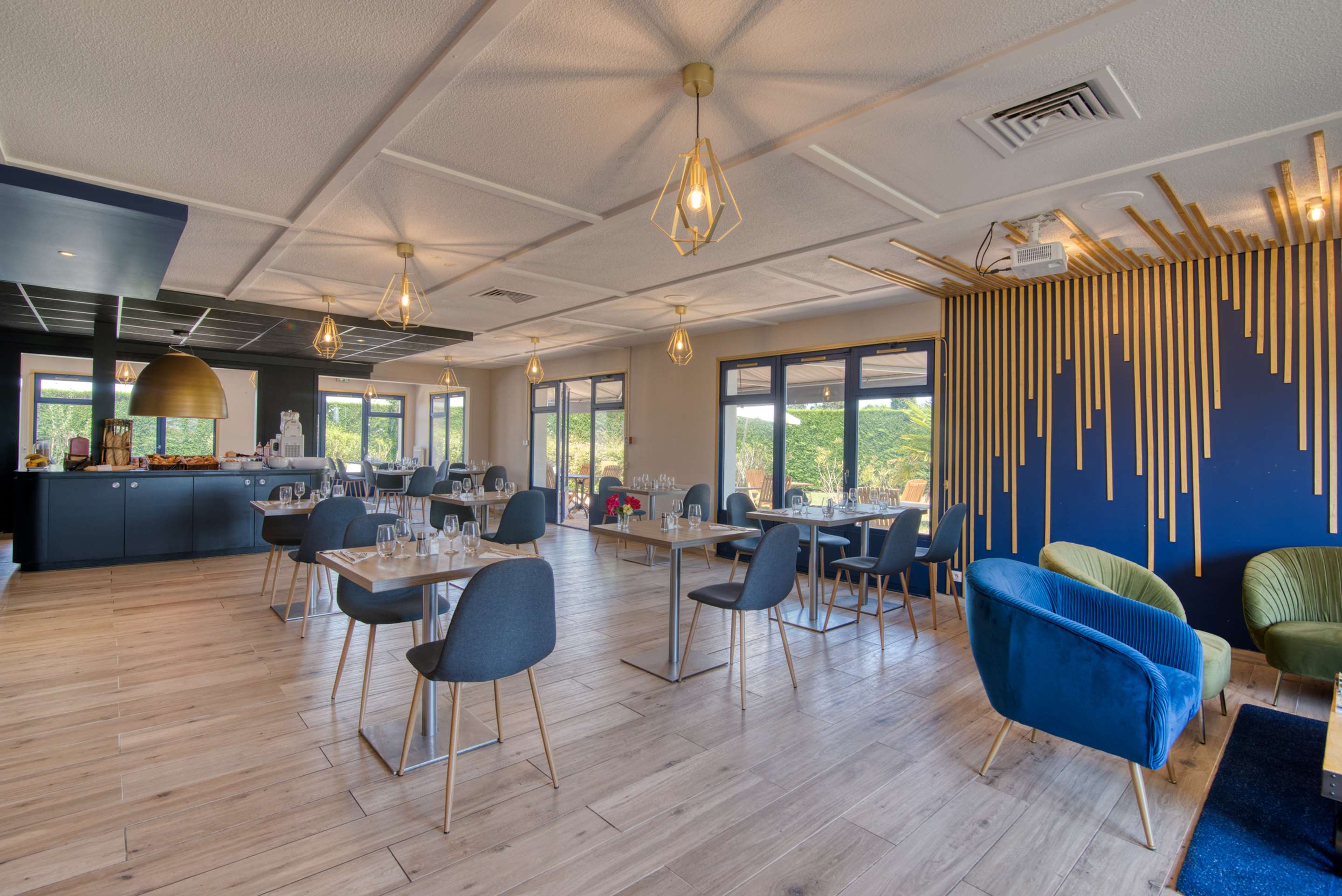Salle du restaurant avec tables dressées, fauteuils en velours bleu et mur décoré de tasseaux dorés